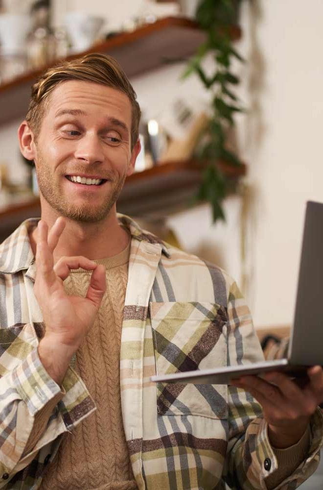 Young man in a plaid shirt smiling and giving an 'okay' gesture while holding a laptop in a cozy cafe setting, showcasing a friendly and engaging atmosphere for remote work or online meetings.